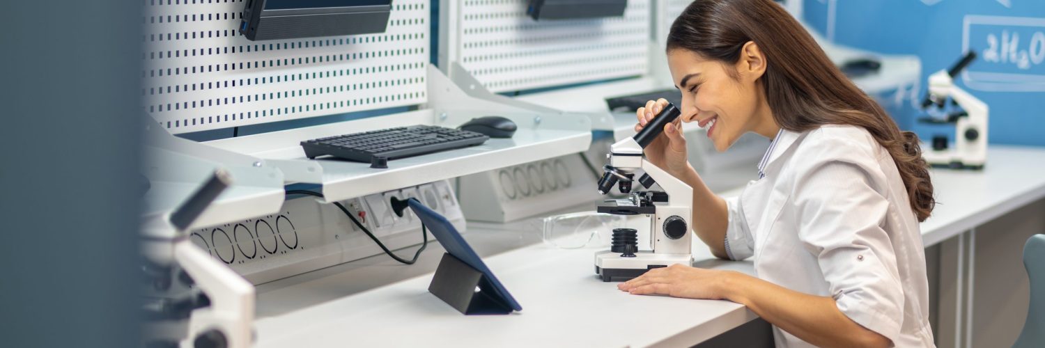 Professionalism. Cheerful woman in white coat sitting at table in chemistry room working with microscope